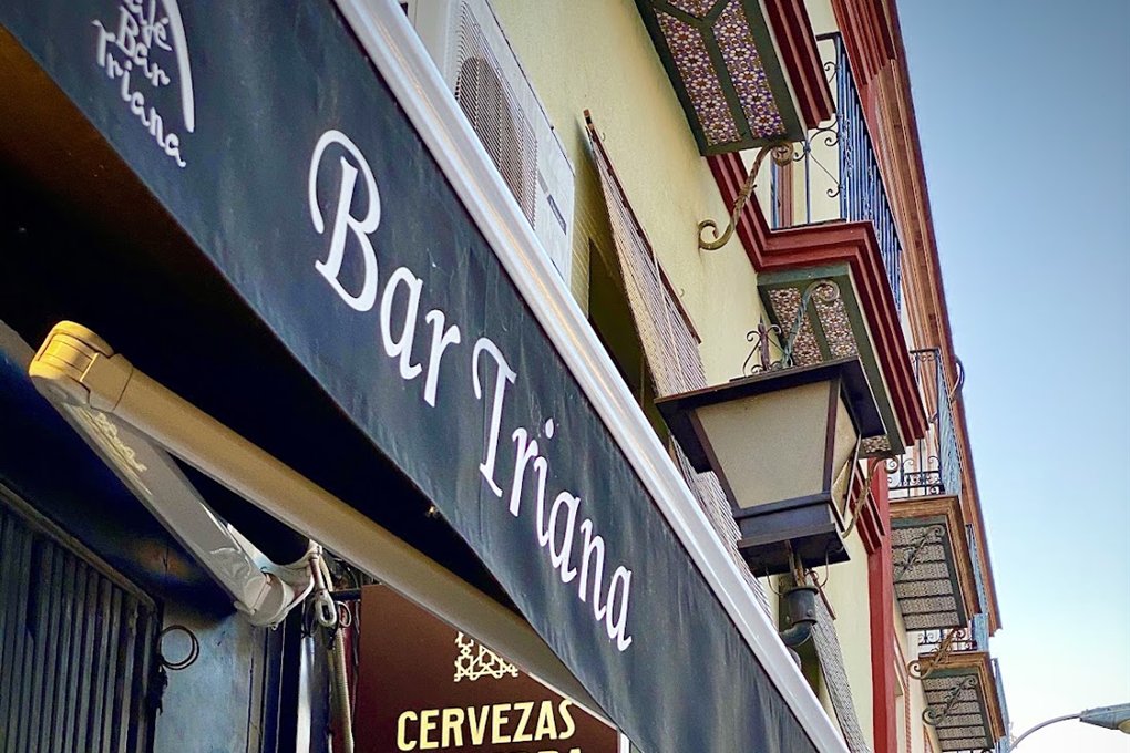 Toldo oscuro con el nombre Bar Triana en letra blanca y el cartel de cervezas, en una calle de fachadas con azulejos